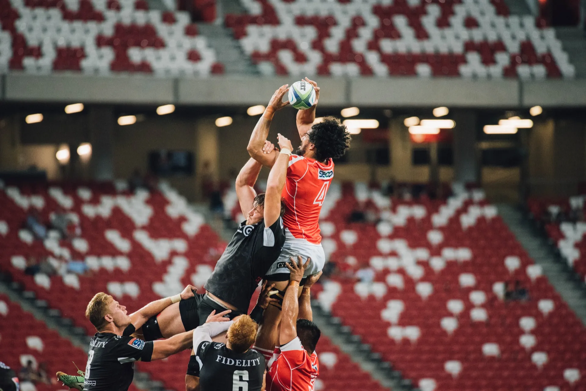 Two rugby players lifted high at a lineout, one catching the ball above the other, in front of a red and white stadium stand