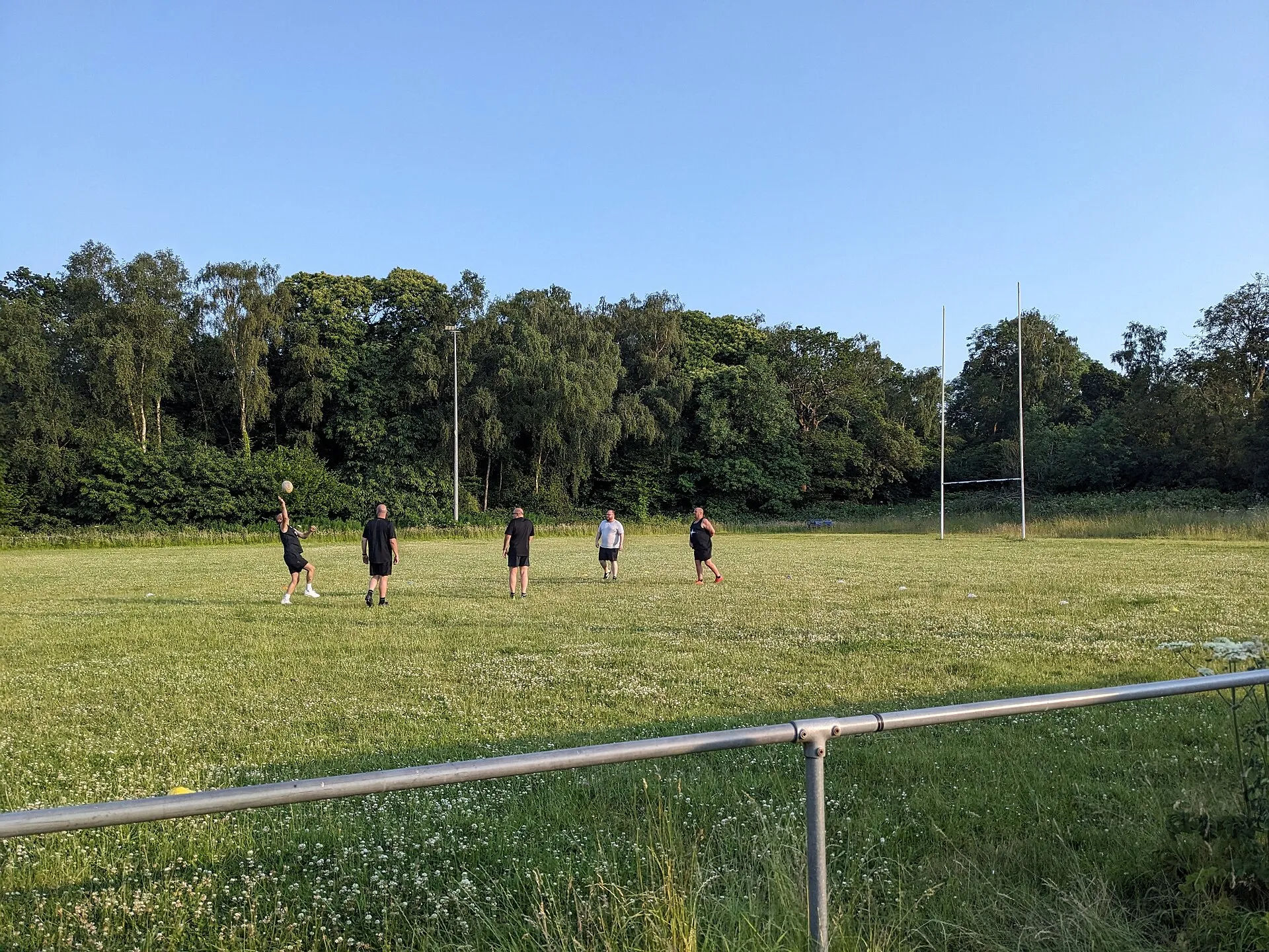Five players passing a ball at an evening training session on an amateur rugby pitch, with the posts and trees behind them