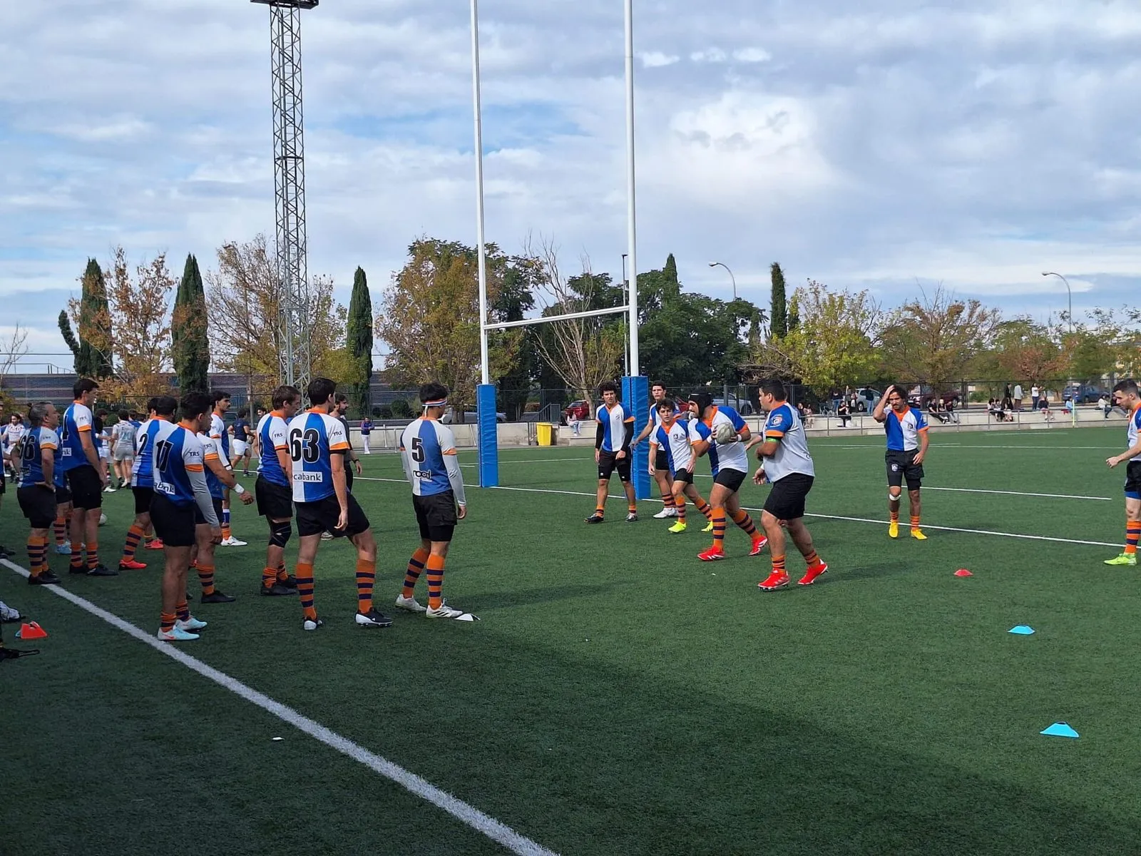 An amateur rugby squad in mixed training kit stands among cones on an artificial pitch before a session