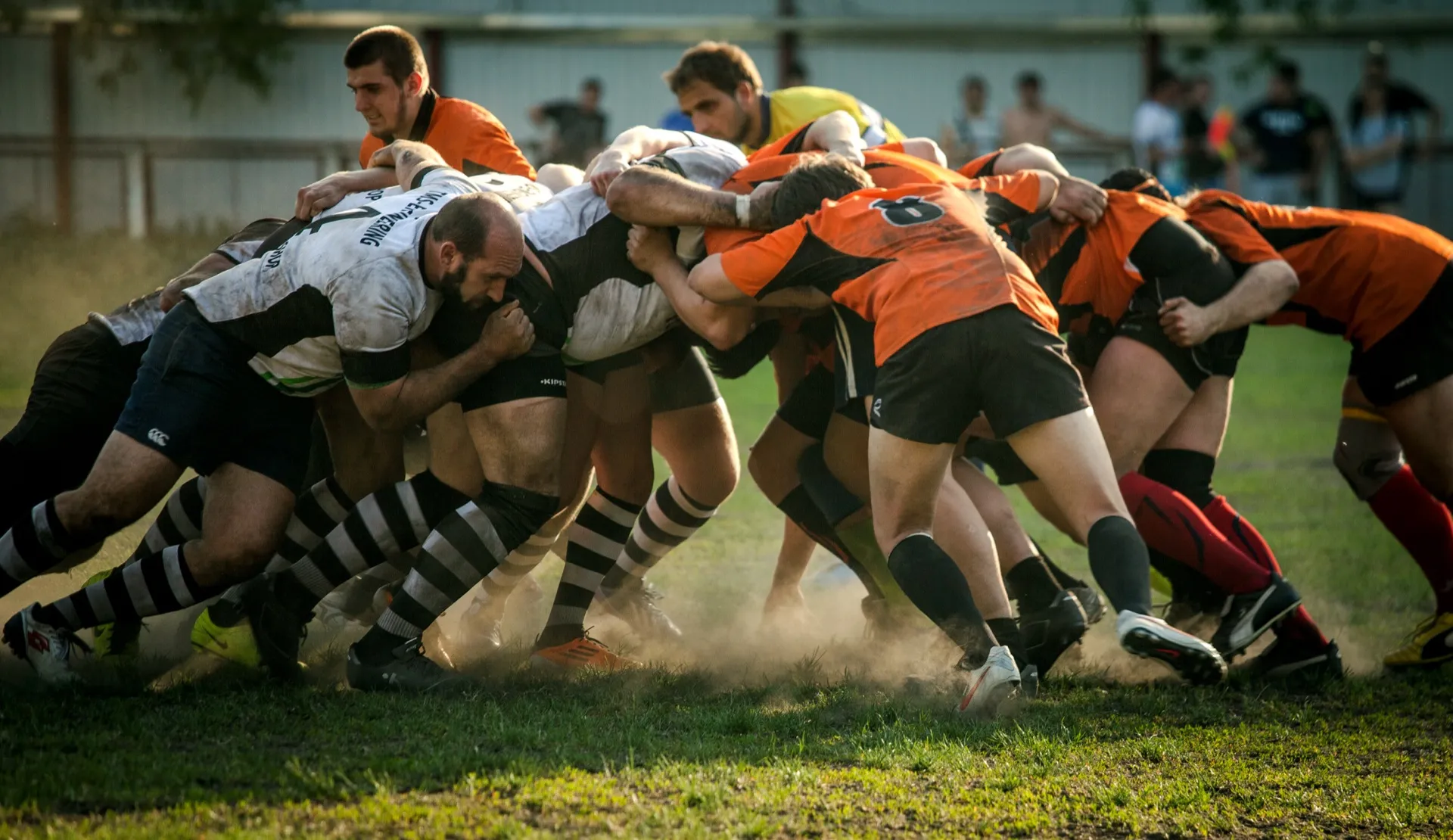 Two amateur rugby packs locked in a scrum on a dusty pitch, one team in orange and black, the other in white