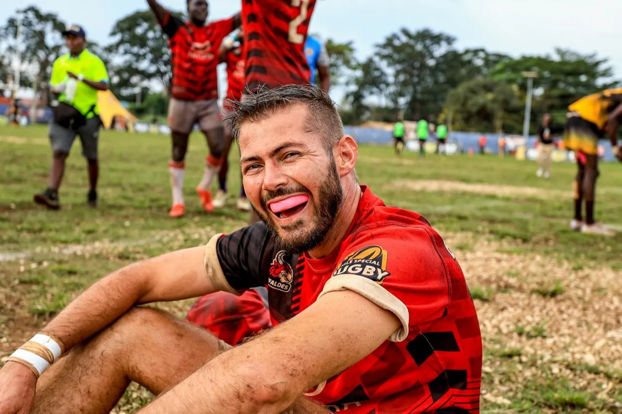 A smiling rugby player in a sponsored shirt sitting on the pitch after a match