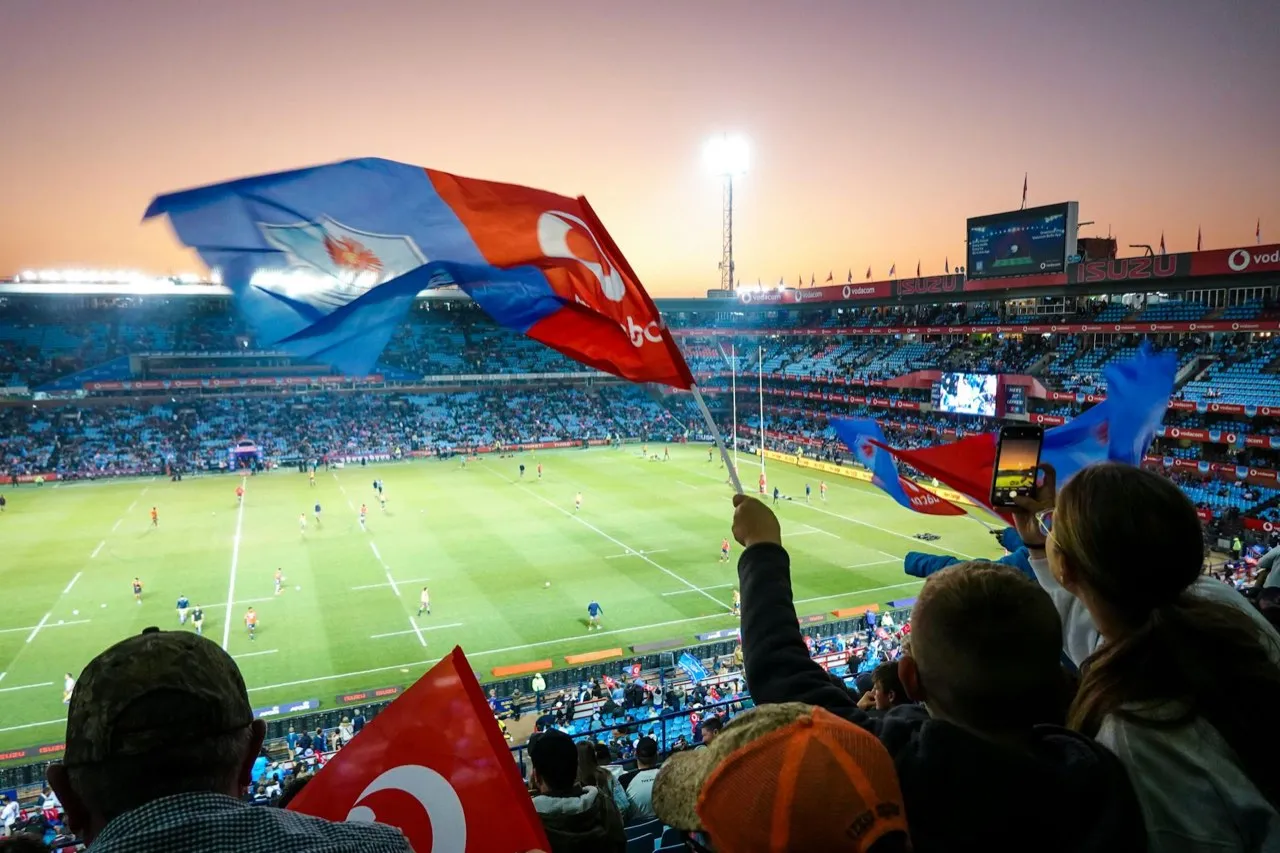 Rugby supporters waving flags in a floodlit stadium at dusk, one filming the match on a phone