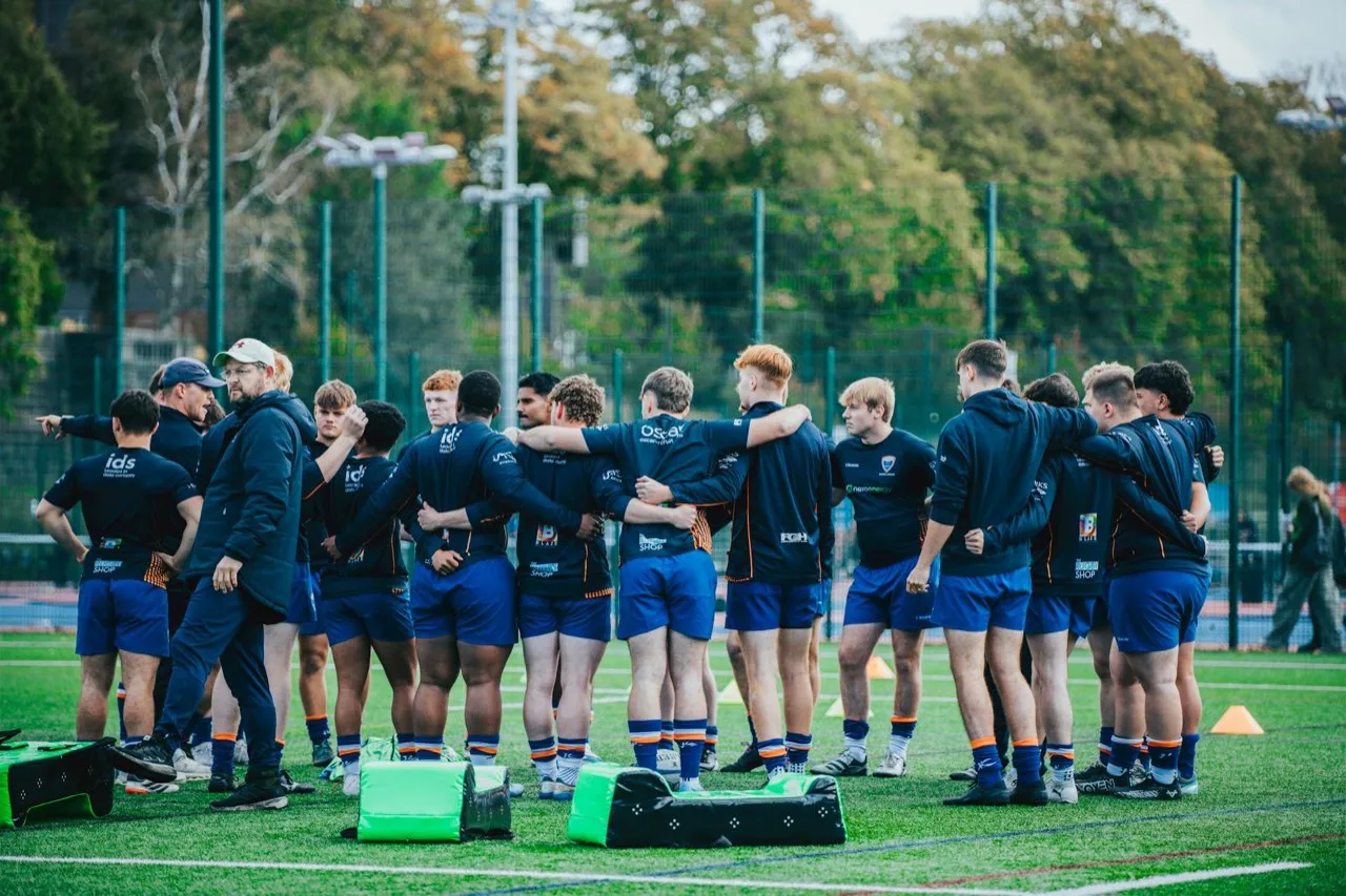 A rugby coach pointing as he talks to a squad of players huddled together on a training pitch