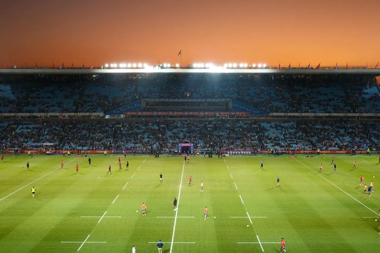 A rugby pitch viewed from the stands at dusk, players warming up under floodlights before kickoff