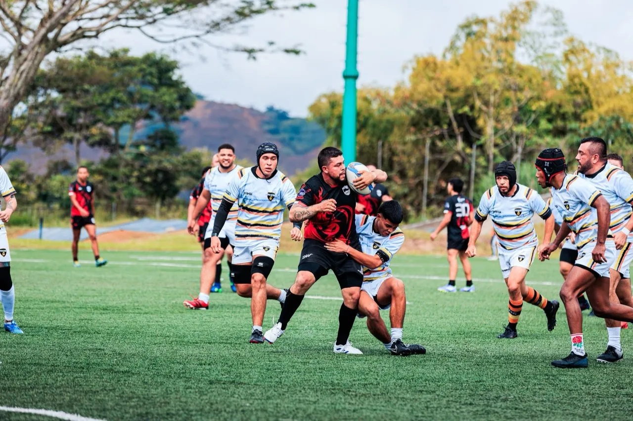 A rugby ball-carrier in a red shirt breaking through a tackle during a match, players around him on the pitch