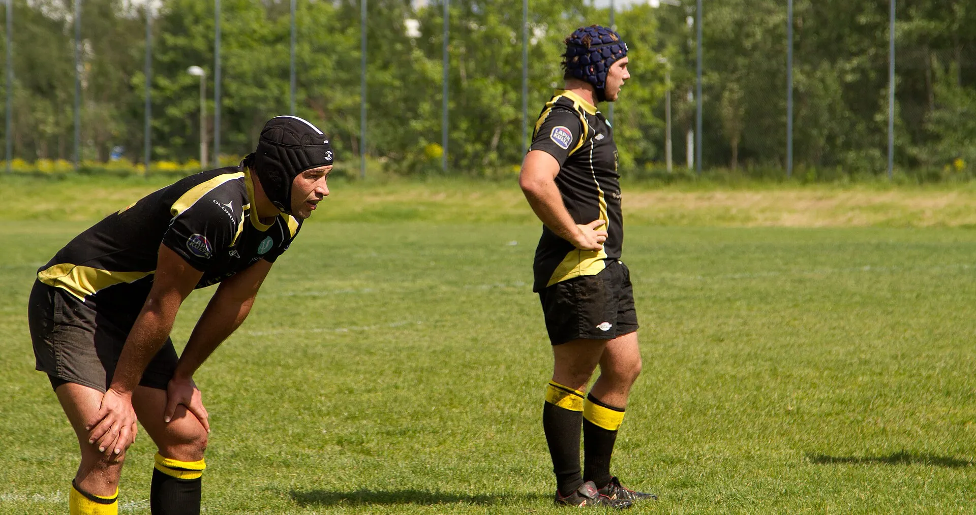 Two rugby players in scrum caps pause and study the play in front of them