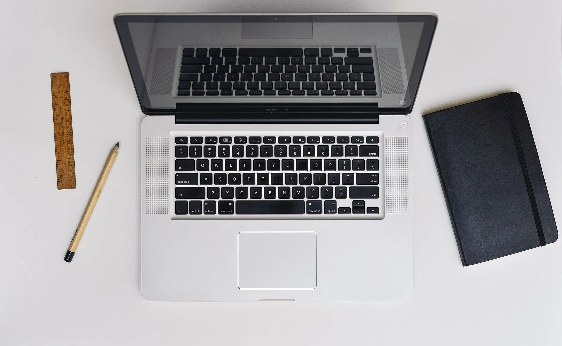 A MacBook Pro seen from above on a white desk, next to a notebook, a pencil and a ruler