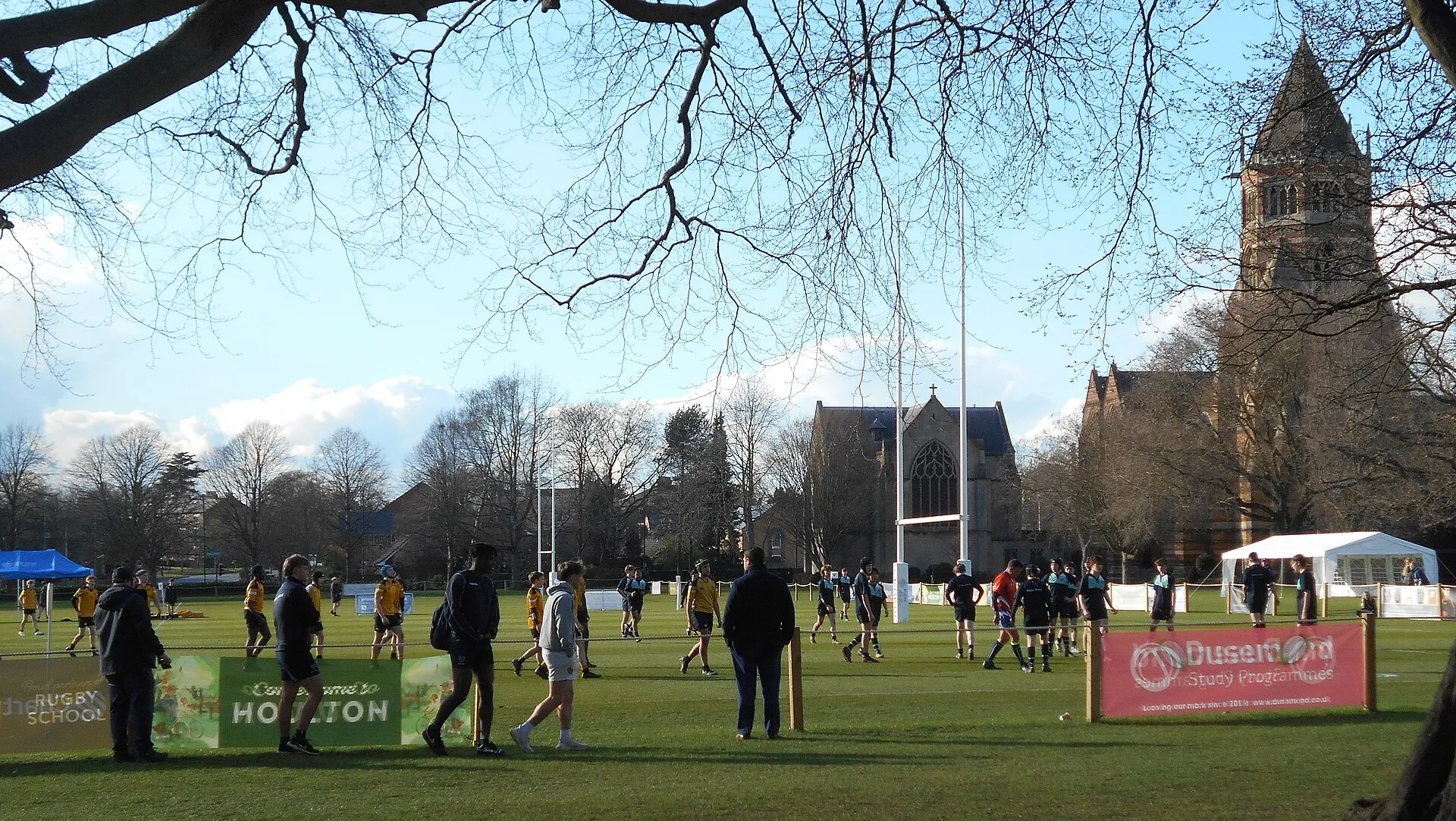 School rugby teams warming up on a playing field in spring sunshine, with a Victorian chapel behind the posts