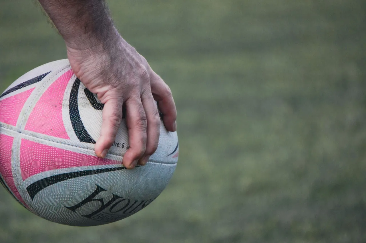 A hand gripping a pink-and-white rugby ball on a grass pitch