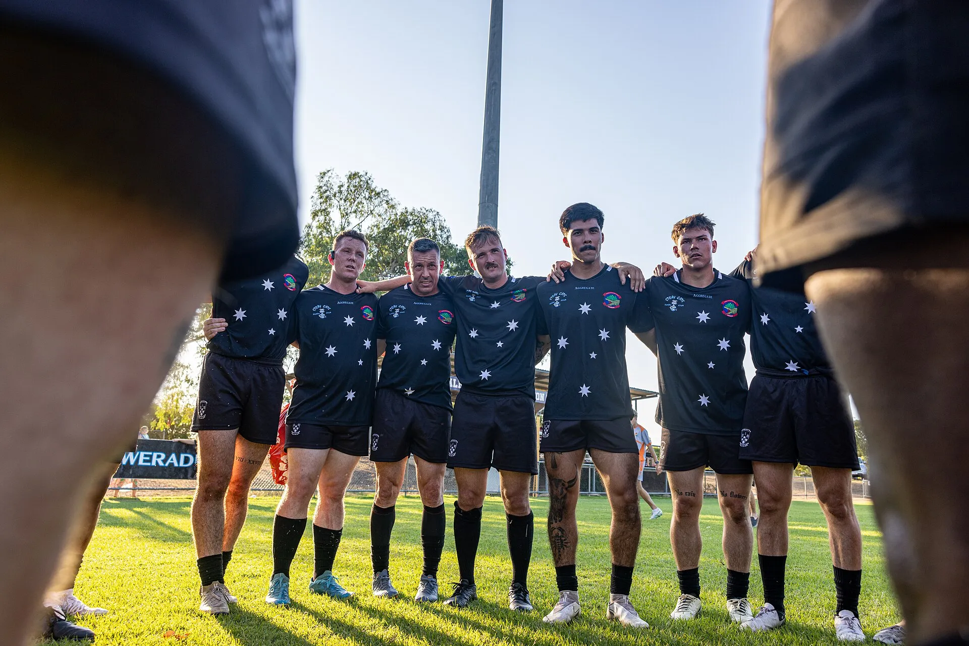 Club rugby players stand arm in arm in a huddle in low evening sun