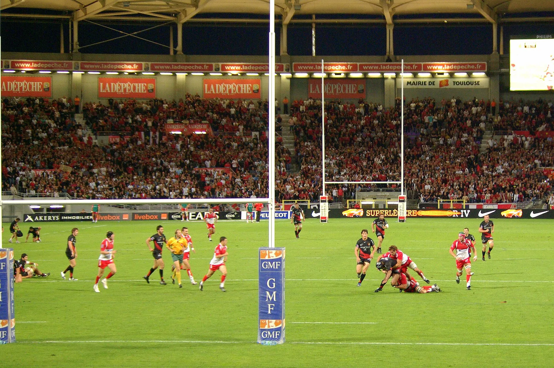A professional rugby match under floodlights in a packed stadium, with a tackle going in near the posts