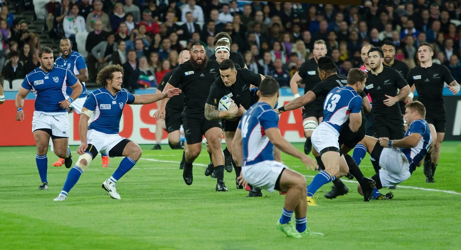 A ball carrier in black runs at a line of defenders in blue and white during an international match
