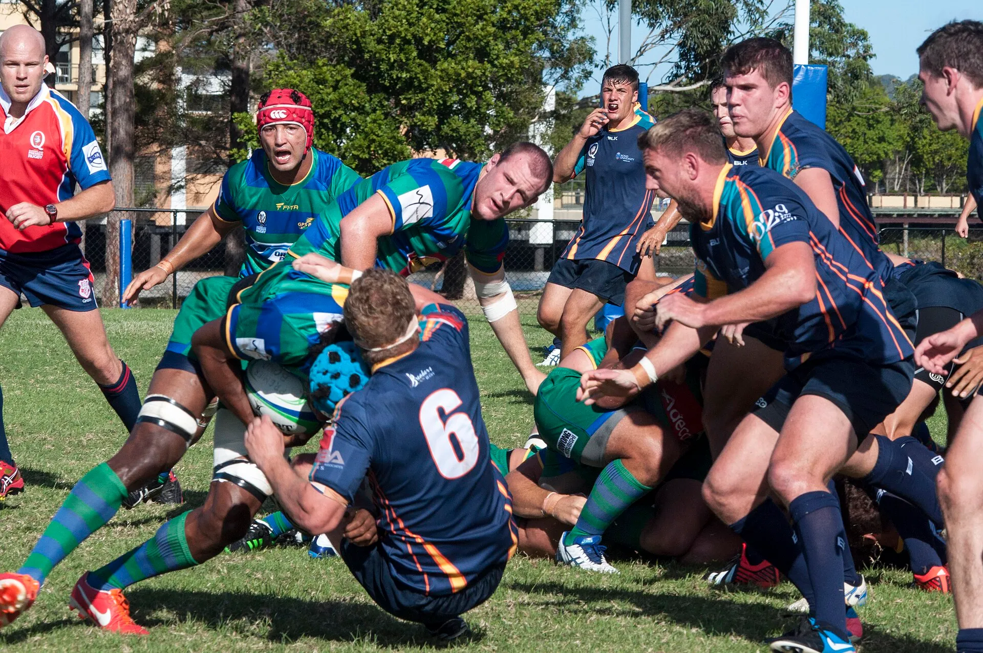A player in the number 6 shirt tackles a ball carrier in an amateur rugby match while the referee looks on