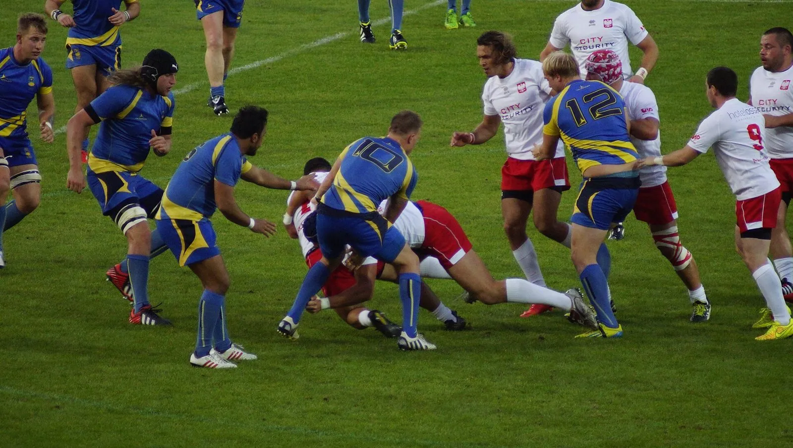 Defenders in blue and yellow close in as a player in white and red is brought to ground in a tackle