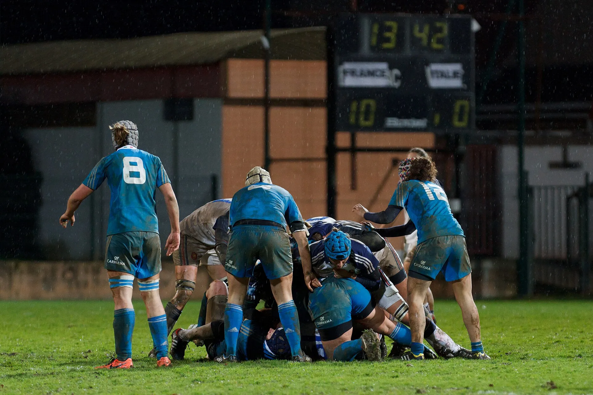 Players pile into a ruck in the rain during a women's international, the scoreboard lit behind them