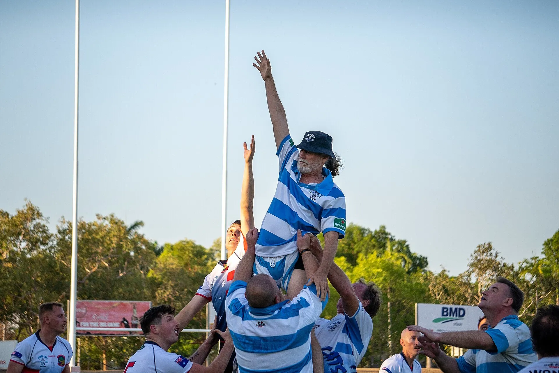 A lineout jumper is lifted to take the throw at an amateur rugby match under a clear sky